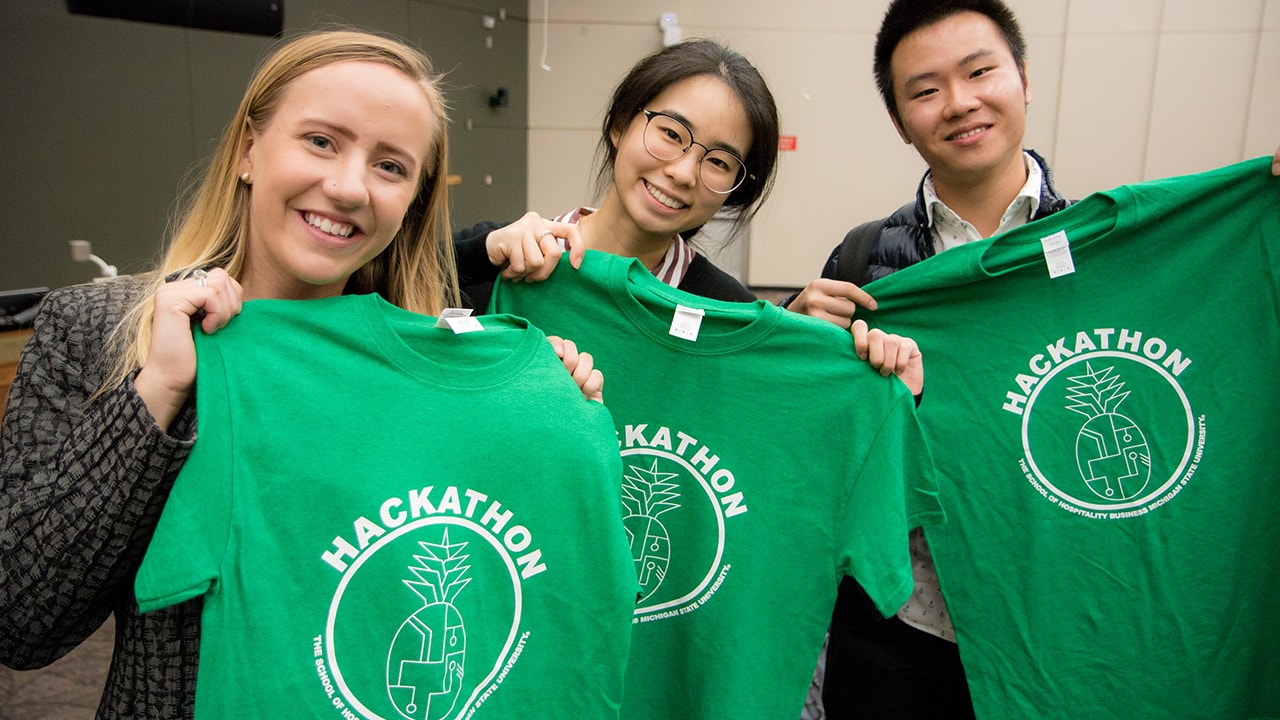 Three Michigan State University students pose in a Broad College lecture hall with green MSU Hilton Hackathon shirts that were given to each of the event's participants. 