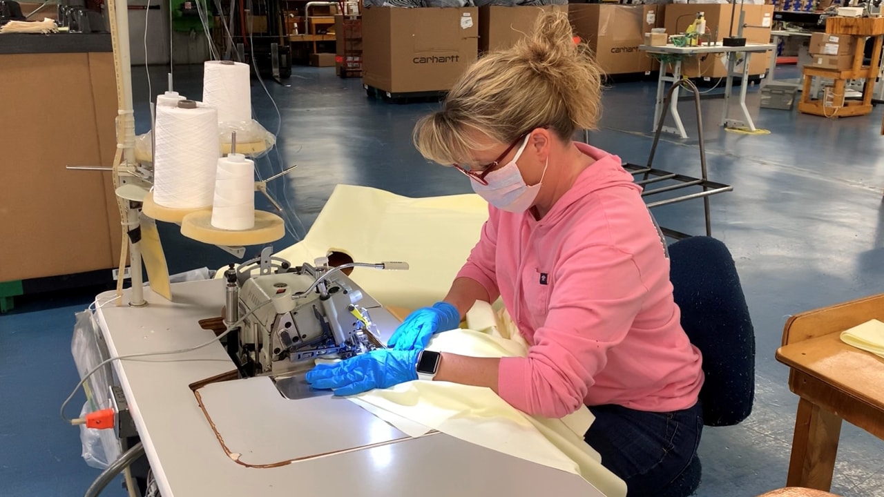 A female Carhartt employee seen at a work station sewing together a medical gown, wearing gloves and a face mask.