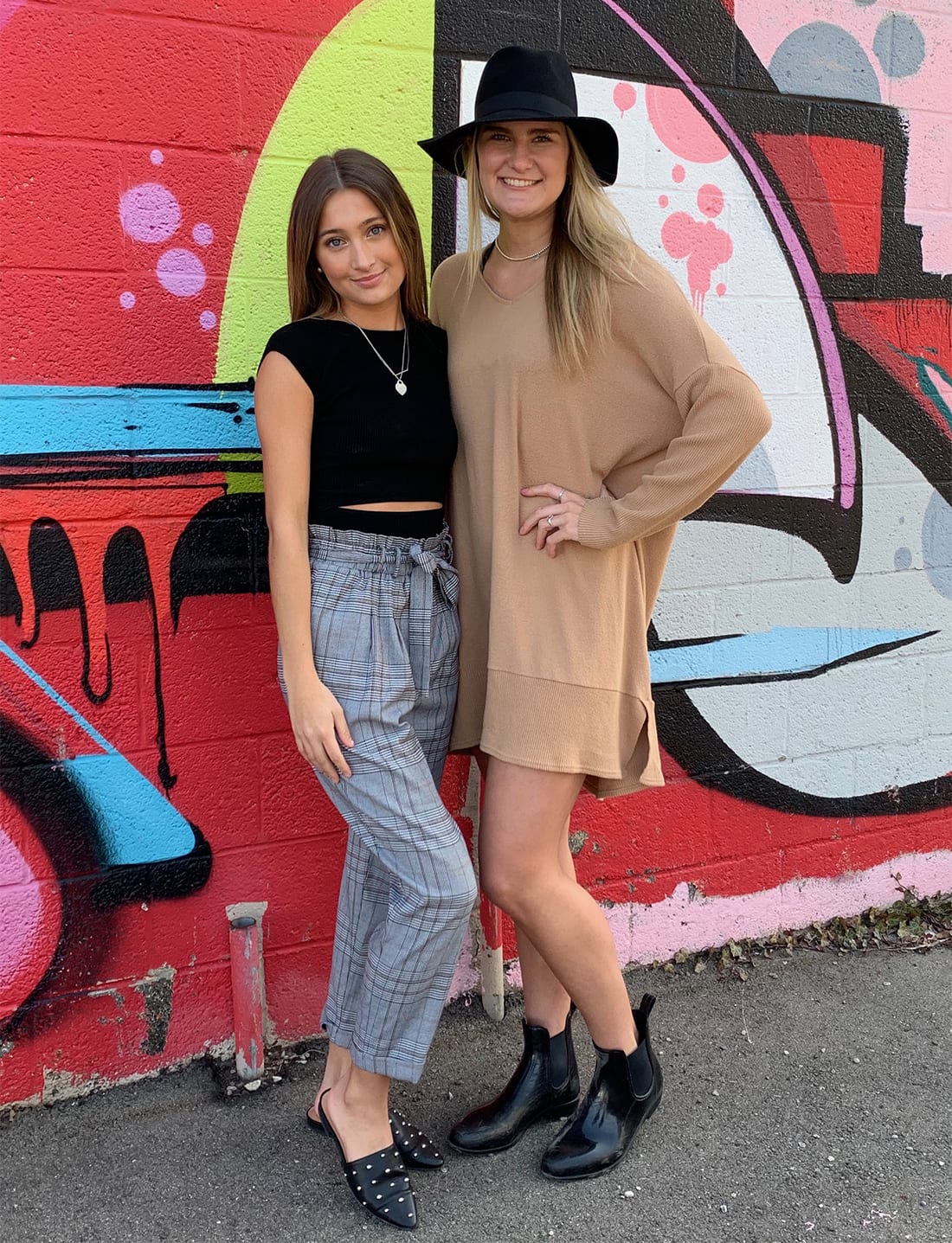 Broad students Jackie Smythe and Olivia Miller stand together in front of a brightly painted brick wall.